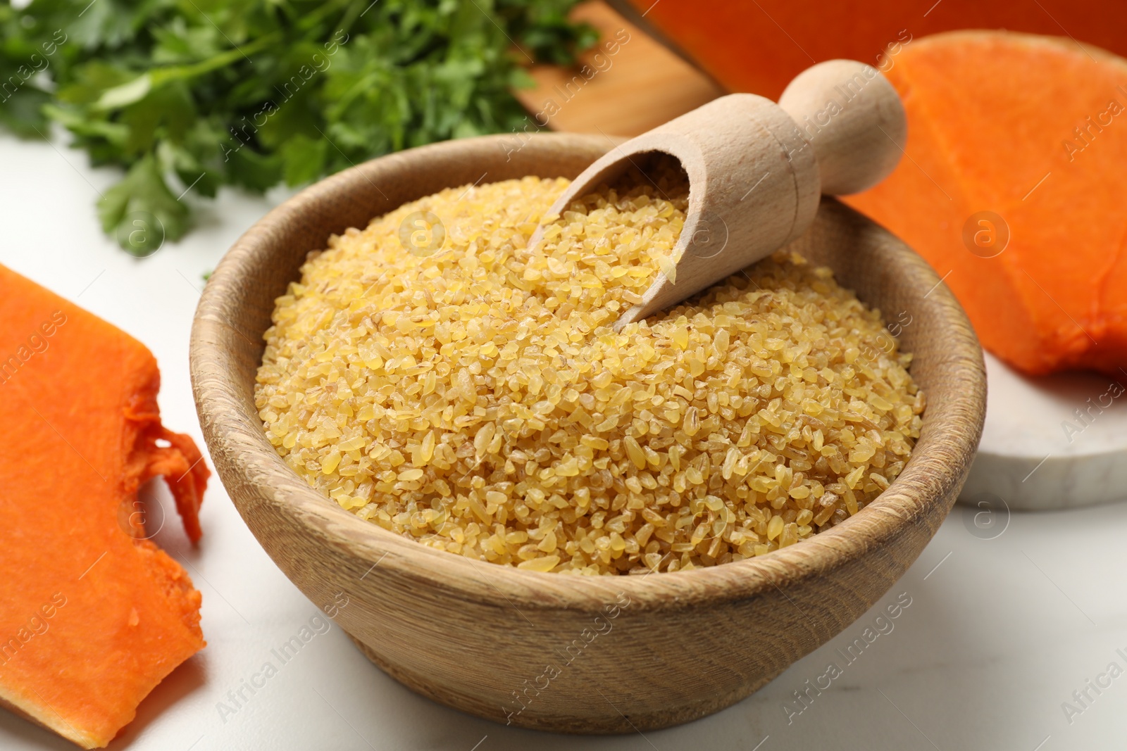 Photo of Raw bulgur in bowl, scoop and pieces of pumpkin on white table, closeup