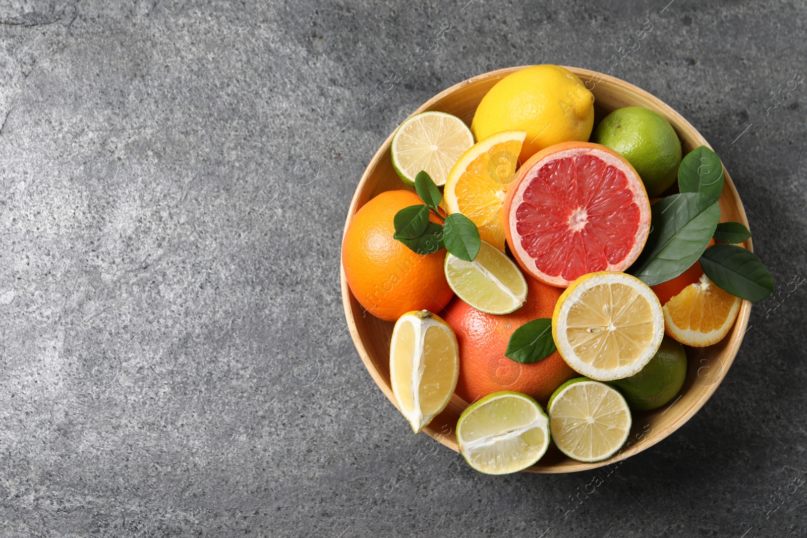 Photo of Different fresh citrus fruits and leaves in bowl on grey textured table, top view. Space for text