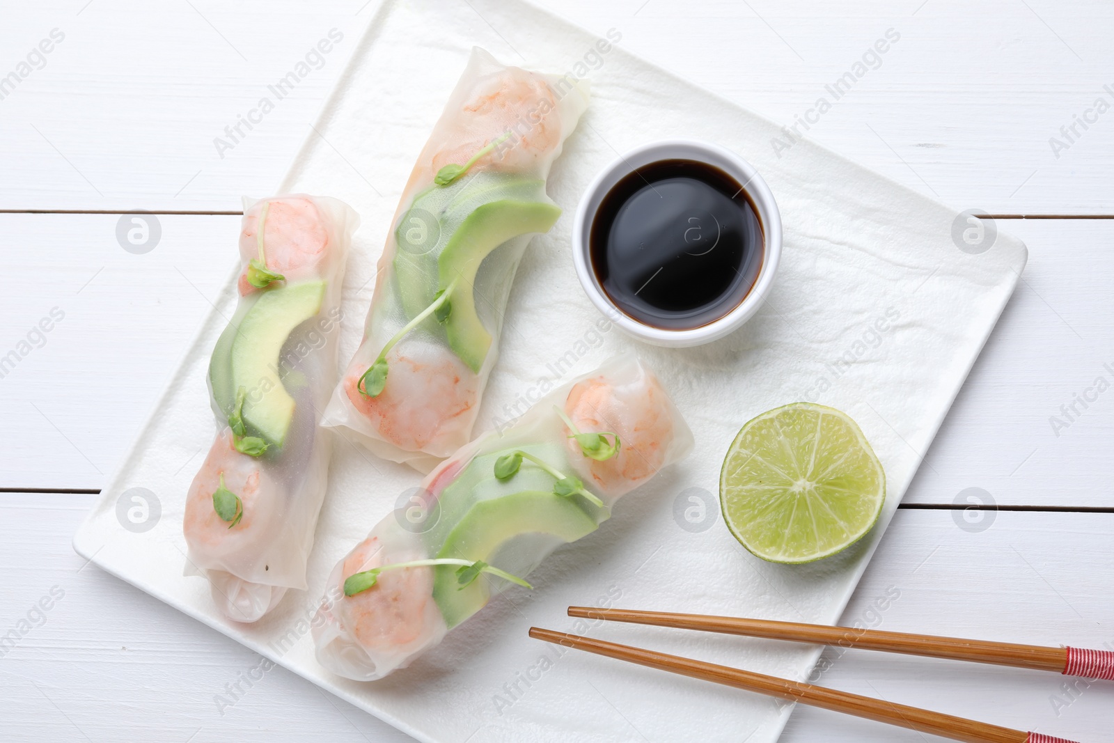 Photo of Delicious spring rolls, lime, soy sauce and chopsticks on white wooden table, top view