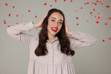 Beautiful young woman under falling heart shaped confetti on grey background
