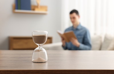 Photo of Hourglass with flowing sand on desk. Man reading book in room, selective focus