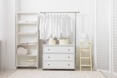 Photo of Wardrobe organization. Rack with different stylish clothes, chest of drawers and shelving unit near white wall indoors