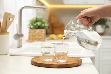 Photo of Woman pouring water from jug into glass at white table in kitchen, closeup