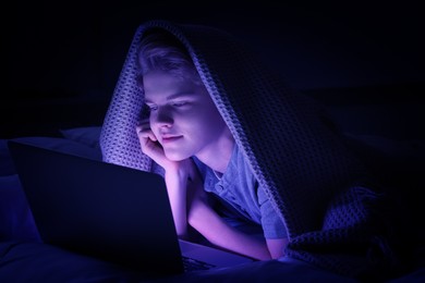 Internet addiction. Teenage boy using laptop under blanket on bed at night. Toned in blue