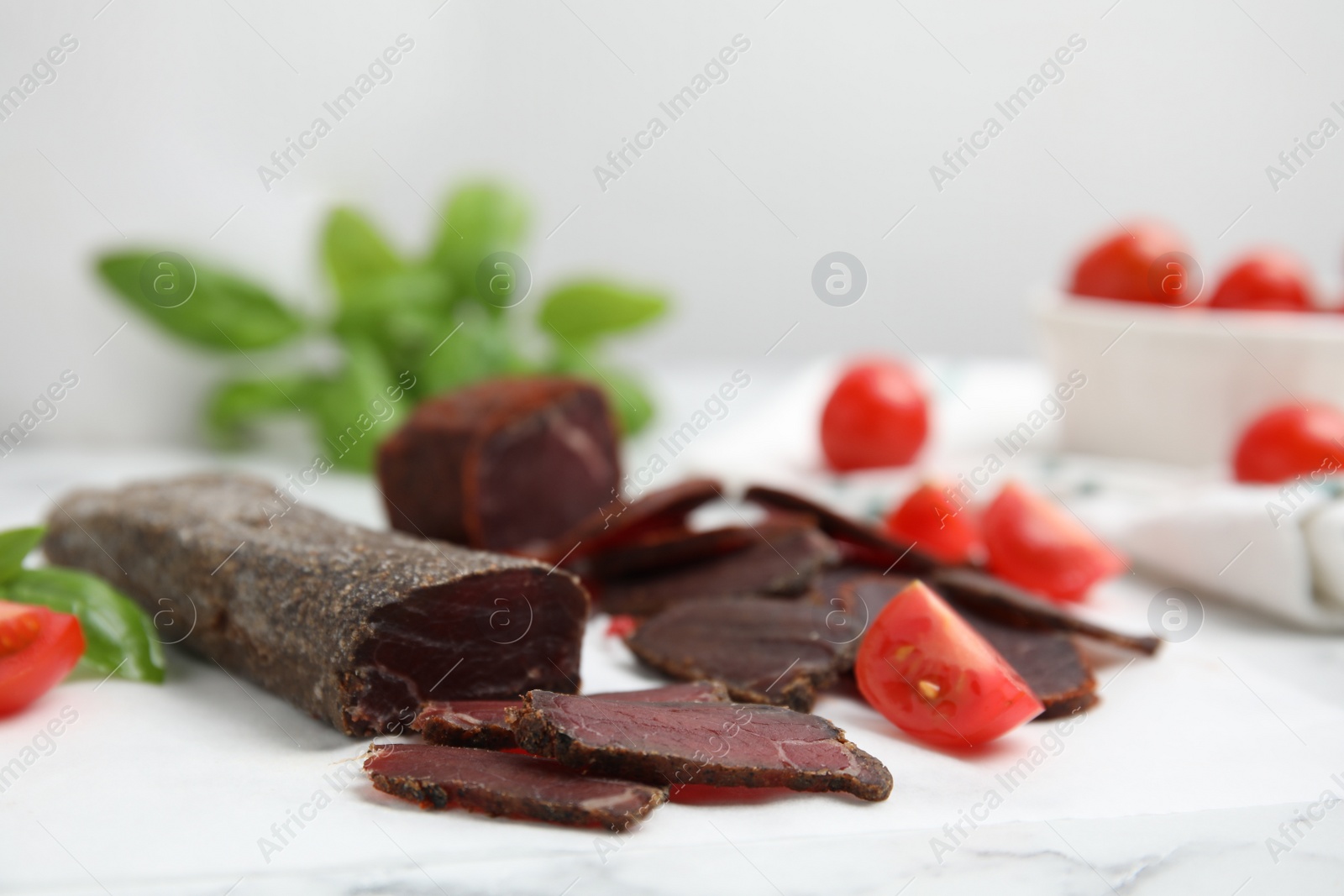 Photo of Delicious dry-cured beef basturma with basil and tomatoes on white marble table, closeup