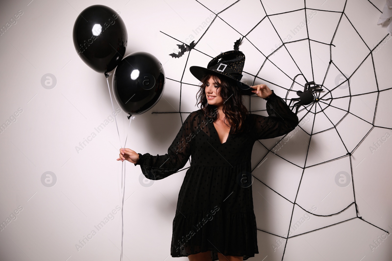 Photo of Woman in witch hat with balloons posing near white wall decorated for Halloween