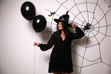 Woman in witch hat with balloons posing near white wall decorated for Halloween