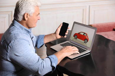 Photo of Man using laptop and phone to buy car at table indoors