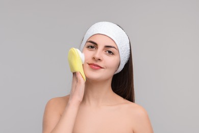 Photo of Young woman with headband washing her face using sponge on light grey background