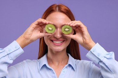 Photo of Happy woman covering eyes with halves of fresh kiwi on violet background