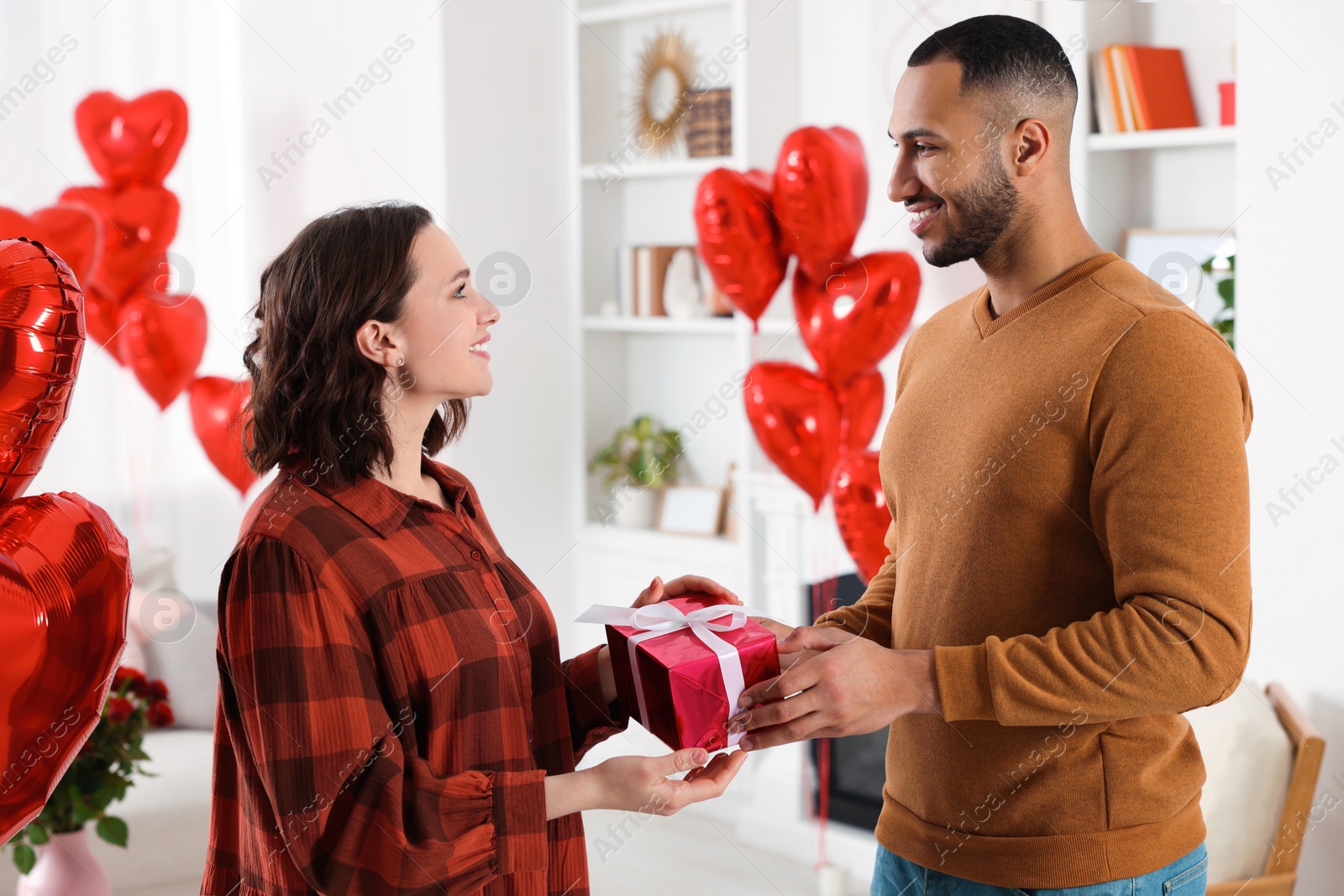 Photo of Lovely couple holding beautiful gift box in room decorated with heart shaped air balloons. Valentine's day celebration