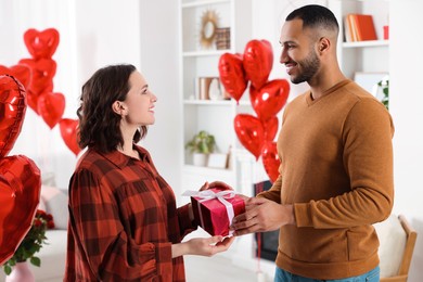 Photo of Lovely couple holding beautiful gift box in room decorated with heart shaped air balloons. Valentine's day celebration