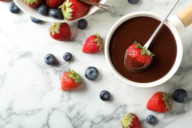 Photo of Dipping strawberry into fondue pot with chocolate on white marble table, flat lay
