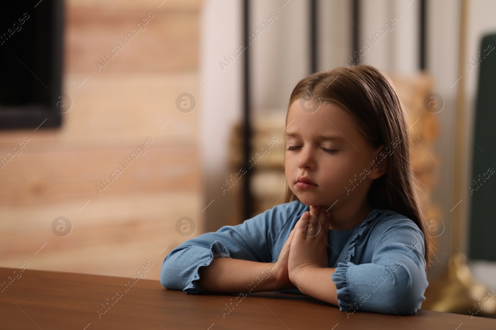 Photo of Cute little girl with hands clasped together praying at table indoors