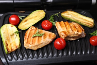 Photo of Electric grill with tasty meat, rosemary and vegetables on table, closeup