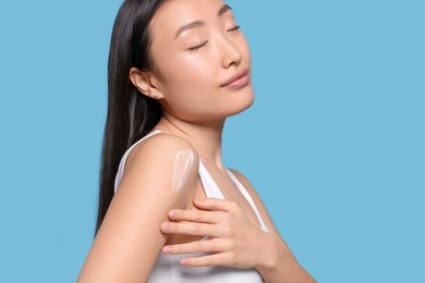 Photo of Beautiful young Asian woman applying body cream onto shoulder on light blue background, space for text