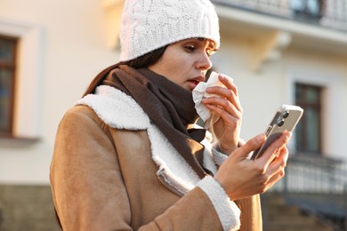 Photo of Woman with tissue blowing runny nose while using phone outdoors. Cold symptom