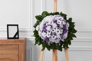 Wreath of violet and white flowers, photo frame with black ribbon in room. Funeral attributes