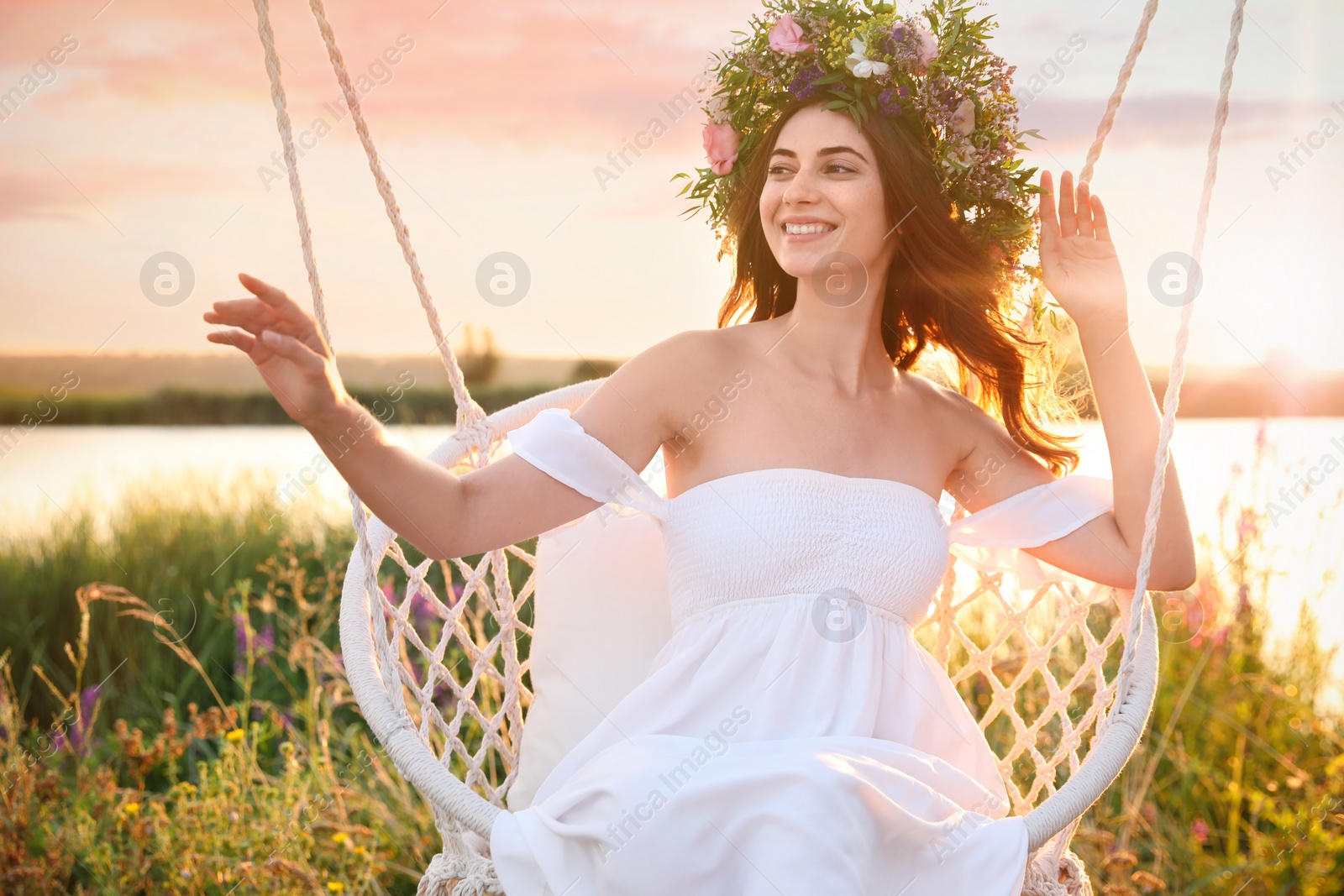 Photo of Young woman wearing wreath made of beautiful flowers on swing chair outdoors at sunset