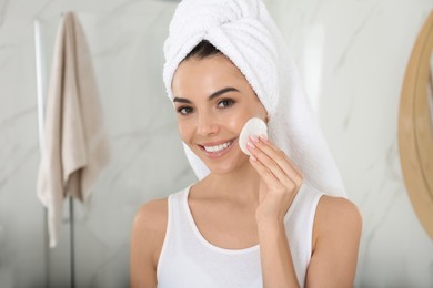 Photo of Happy young woman cleaning face with cotton pad in bathroom