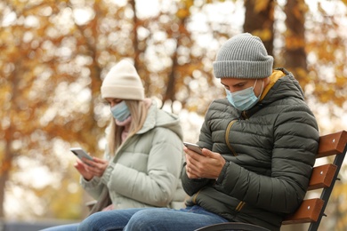 Photo of People in medical masks keeping distance while sitting on bench outdoors. Protective measures during coronavirus quarantine