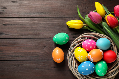 Photo of Colorful Easter eggs in decorative nest and tulips on wooden background, flat lay. Space for text