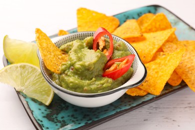 Bowl of delicious guacamole with chili pepper, nachos chips and lime on white wooden table, closeup