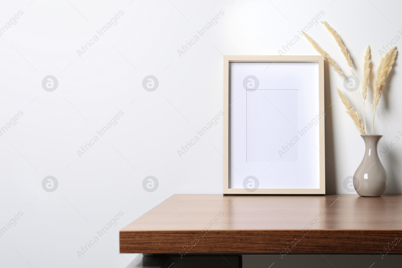 Photo of Empty photo frame and vase with dry decorative spikes on wooden table. Mockup for design