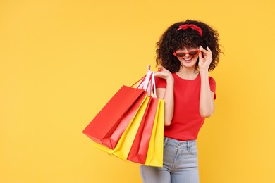 Photo of Happy young woman with shopping bags and stylish sunglasses on yellow background. Space for text