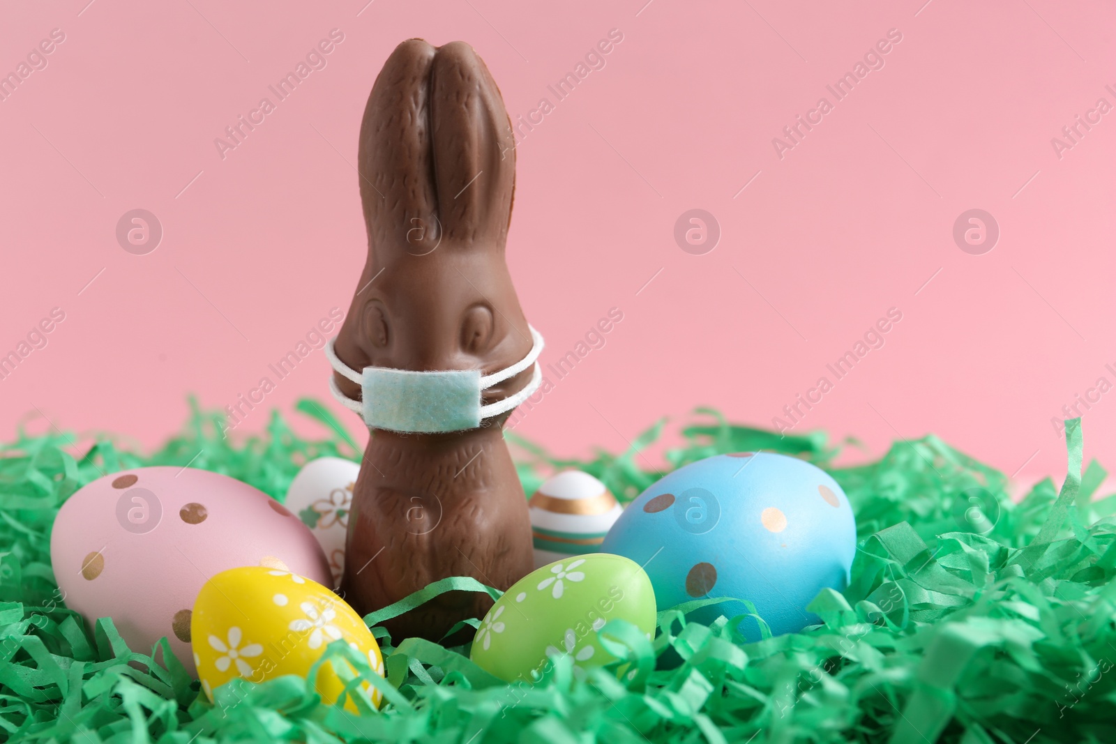 Photo of Chocolate bunny with protective mask and eggs against pink background, space for text. Easter holiday during COVID-19 quarantine