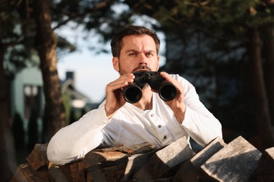 Concept of private life. Curious man with binoculars spying on neighbours over firewood outdoors