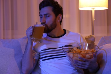 Photo of Man with chips and glass of beer on sofa at night. Bad habit