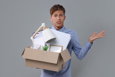 Photo of Confused unemployed young man with box of personal office belongings on grey background