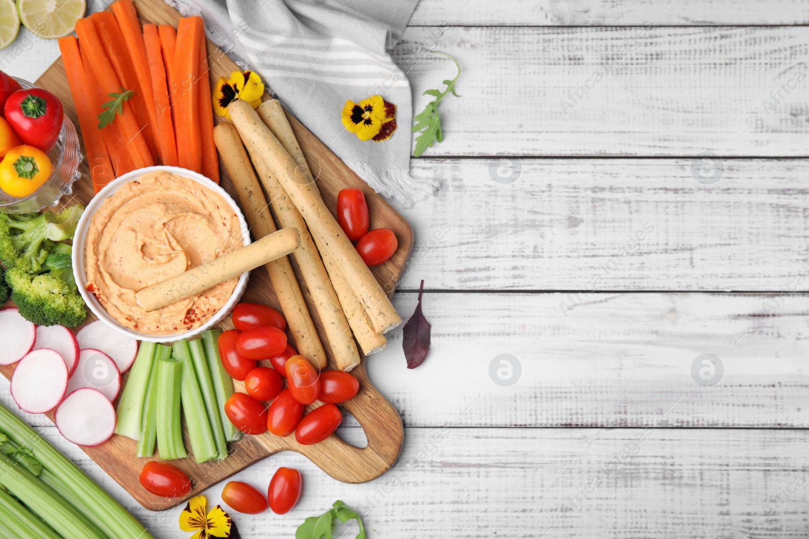 Photo of Board with delicious hummus, grissini sticks and fresh vegetables on white wooden background, flat lay. Space for text