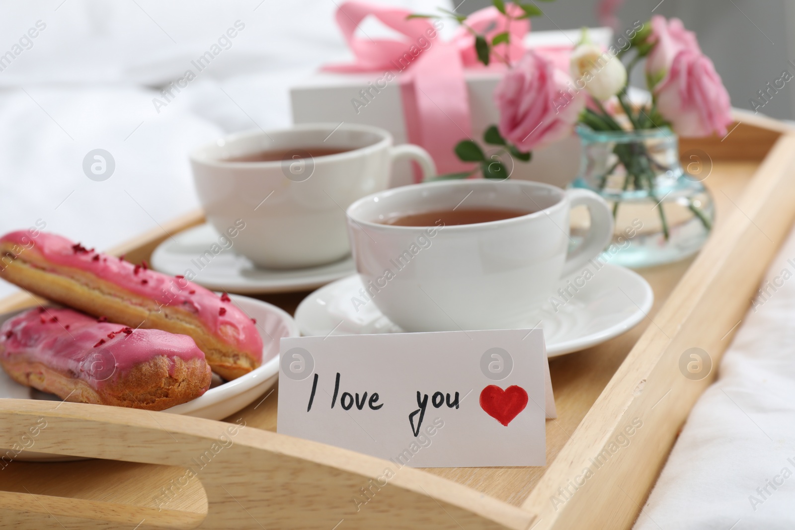 Photo of Tasty breakfast served in bed. Delicious eclairs, tea, gift box, flowers and card with phrase I Love You on tray, closeup