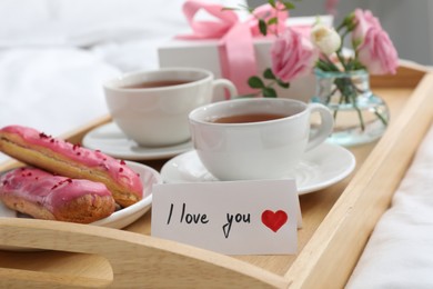 Photo of Tasty breakfast served in bed. Delicious eclairs, tea, gift box, flowers and card with phrase I Love You on tray, closeup