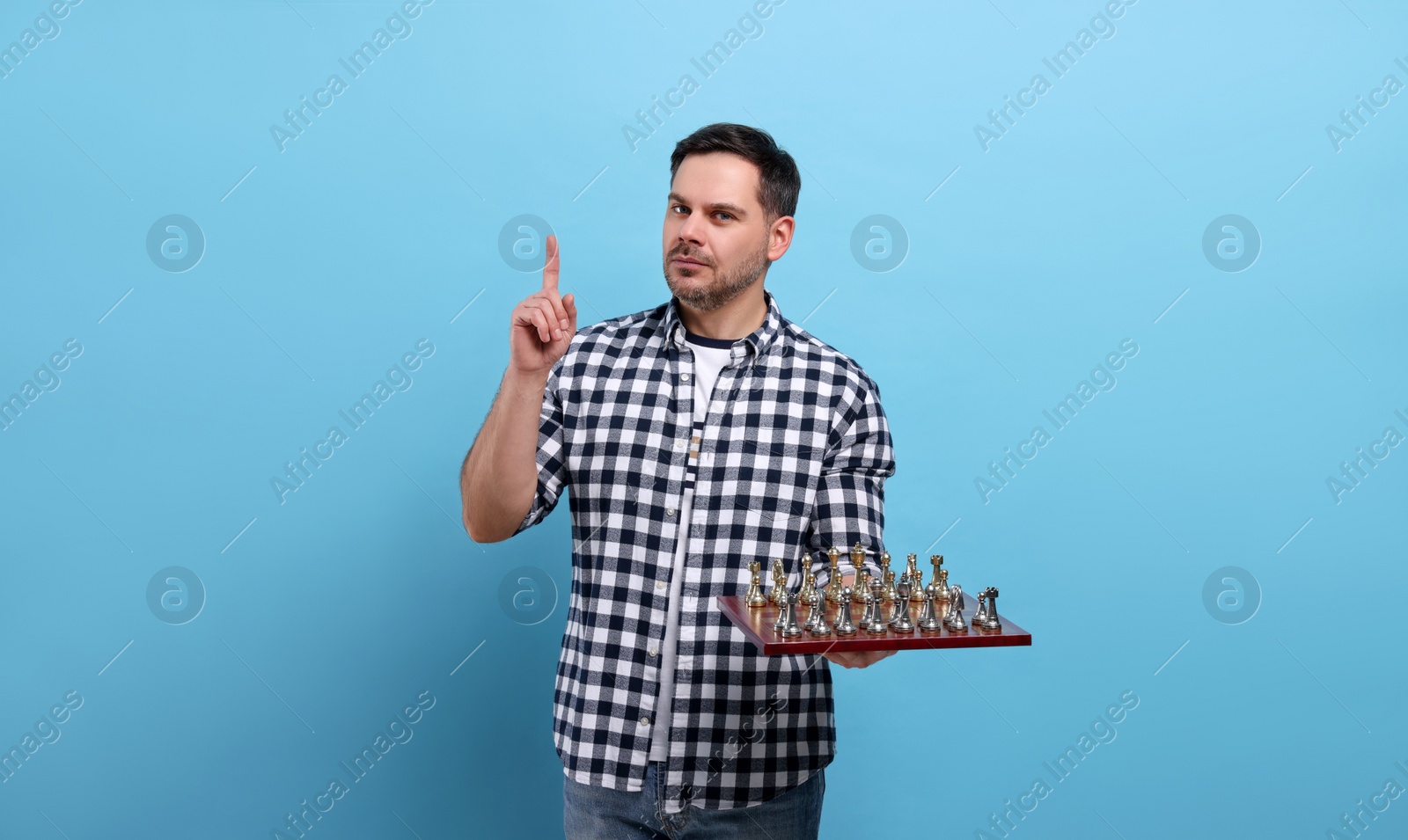 Photo of Adult man holding chessboard with game pieces and pointing up with finger on light blue background