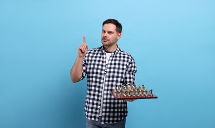 Photo of Adult man holding chessboard with game pieces and pointing up with finger on light blue background