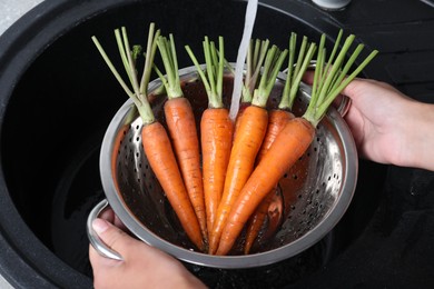 Woman washing ripe carrots in colander with running water over sink, closeup