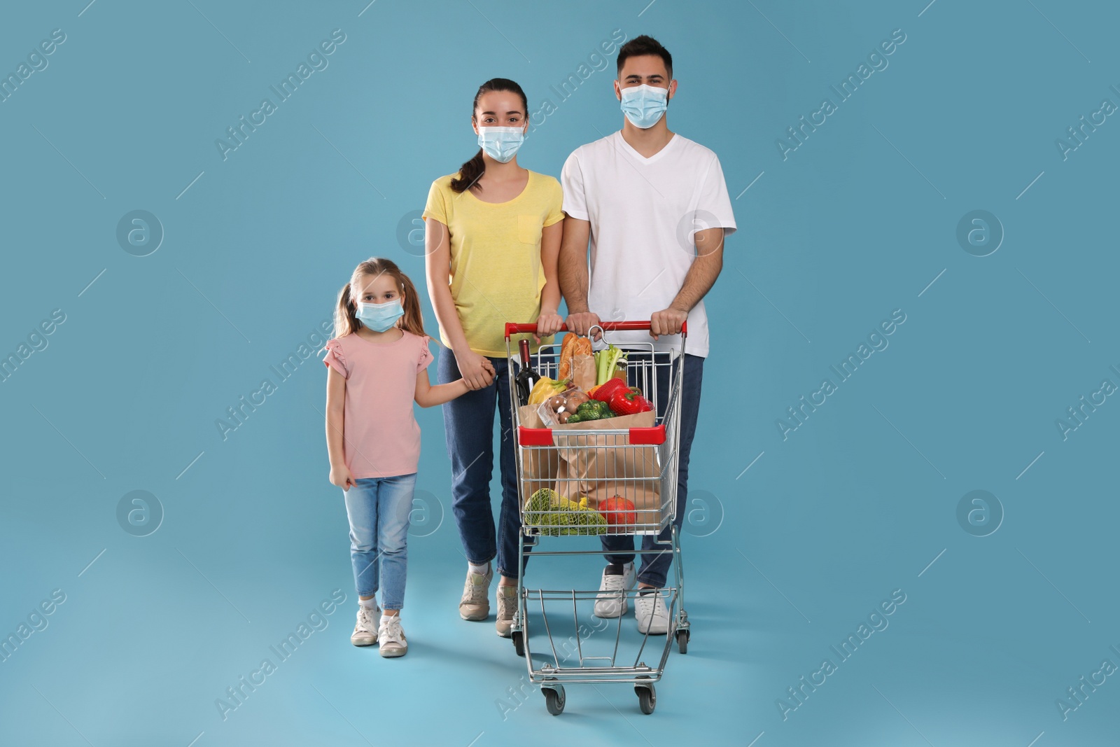 Photo of Family with protective masks and shopping cart full of groceries on light blue background