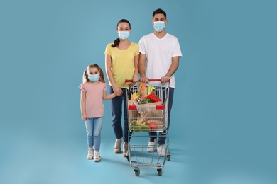 Family with protective masks and shopping cart full of groceries on light blue background