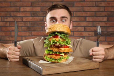 Photo of Young hungry man with cutlery eating huge burger at table
