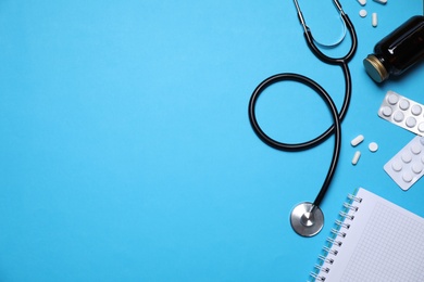 Stethoscope, pills and notebook on light blue background, flat lay. Space for text