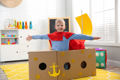 Little child in red cape playing with ship made of cardboard box at home