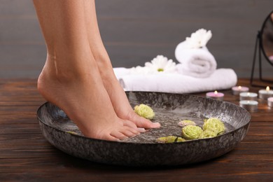 Photo of Woman soaking her feet in bowl with water and flowers on wooden surface, closeup. Pedicure procedure