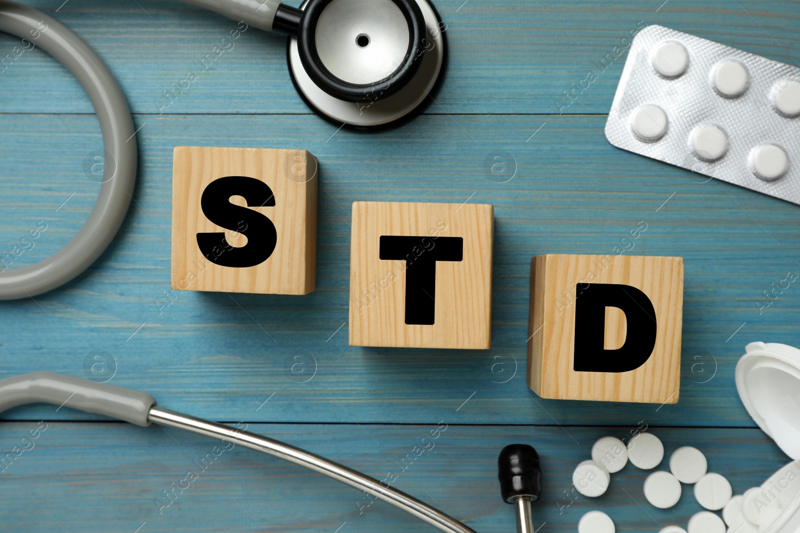 Photo of Abbreviation STD made with cubes, pills and stethoscope on light blue wooden table, flat lay