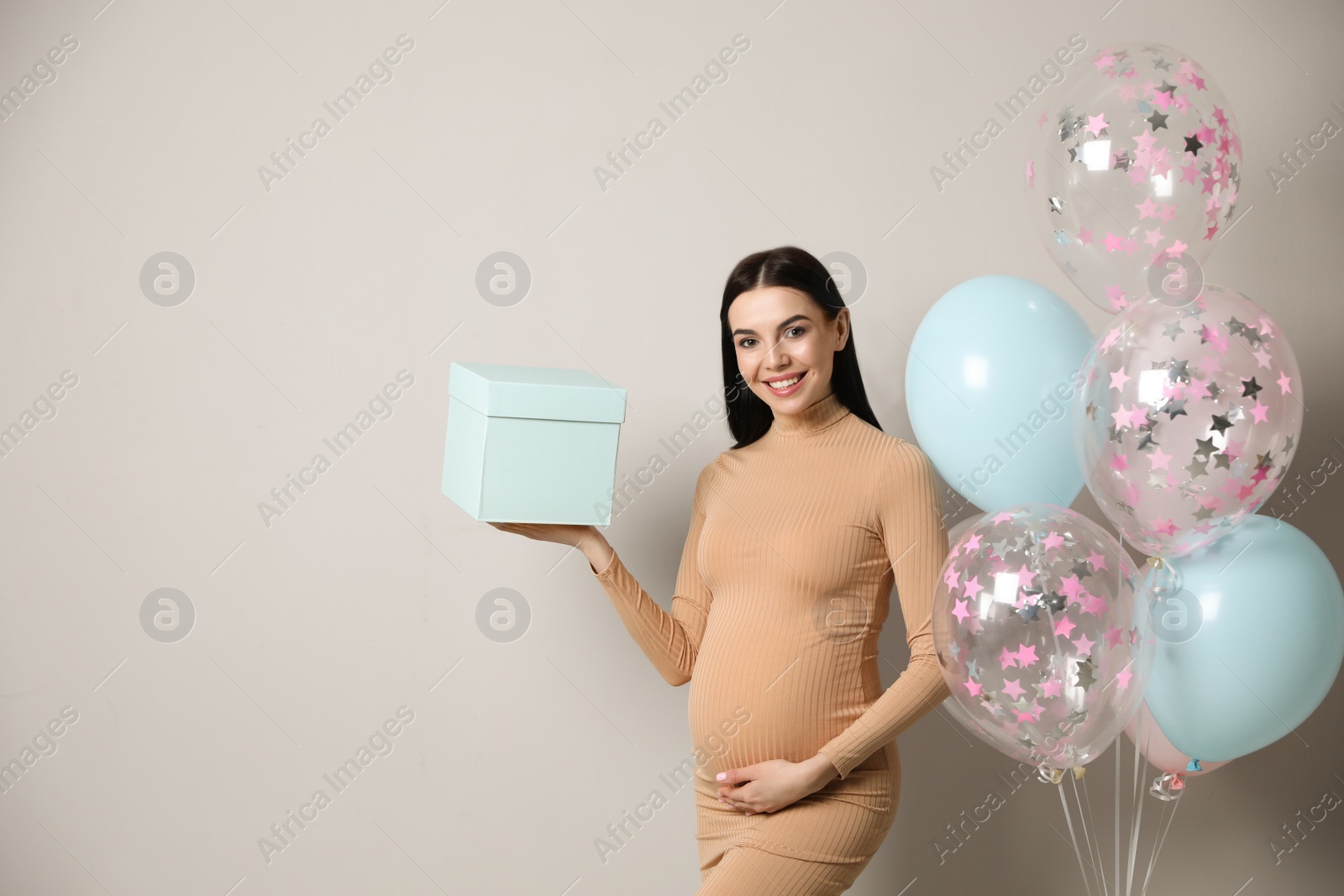 Photo of Happy pregnant woman with gift box and balloons near grey wall, space for text. Baby shower party