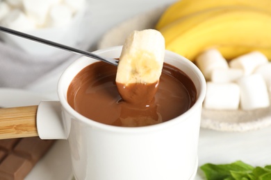 Dipping banana into fondue pot with chocolate on white wooden table, closeup