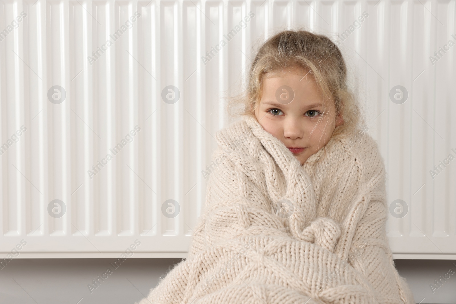 Photo of Little girl covered with knitted plaid sitting near heating radiator at home, space for text