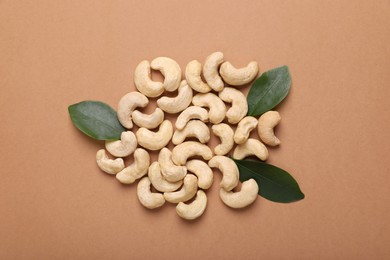 Photo of Pile of tasty cashew nuts and green leaves on pale brown background, flat lay